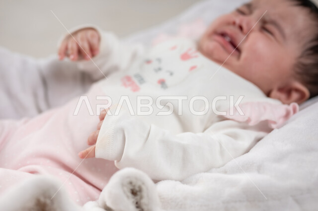 الملامح الناعمة والبريئة، مفهوم النعومة والطفولة، رضيعة سعودية بايماءات البكاء والحاجة إلى شيء ما, بورتريه لطفلة عربيه خليجيه عمانية لطيفة ترتدي لباس قطني مستلقية على كرسي باللون الوردي، خلفية بيضاء