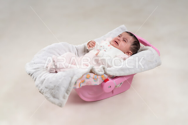ايماءات البكاء والحزن، رضيعة سعودية بحاجة إلى حنان الام, مفهوم النعومة والطفولة، الملامح الناعمة والبريئة، بورتريه لطفلة عربيه خليجيه عمانية لطيفة ترتدي لباس قطني مستلقية على كرسي باللون الوردي، خلفية بيضاء