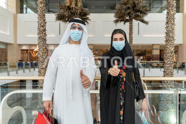 The joy of shopping in malls, adhering to Covid-19 prevention measures, avoiding the spread of infection from the Corona virus, a close-up photo of an Arab Gulf Emirati couple wearing protective masks, carrying colorful paper bags and raising their thumbs up with gestures of acceptance and satisfaction