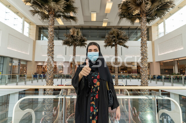تجنب انتشار العدوى من فايروس كورونا, الإلتزام بإجراءات الوقاية من كوفيد - 19, صورة مقربة لإمرأة عربية خليجية اماراتية ترتدي الحجاب والعباءة السوداء والكمامة الواقية ترفع اصبع الإبهام  الى الأعلى بإيماءات القبول والرضا, متعة التسوق في المولات