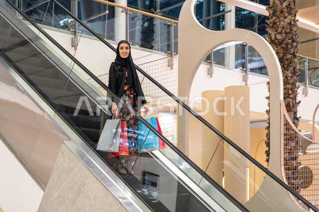 الاستفادة من العروض والخصومات في المول, تعابير السعادة والسرور, الاستمتاع بالشراء وانتقاء المستلزمات والهدايا، الانتقال والنزول باستخدام السلم الكهربائي, إمرأة عربية خليجية اماراتية ترتدي الحجاب والعباءة تتجول في مركز التسوق تحمل الأكياس الورقية الملونة