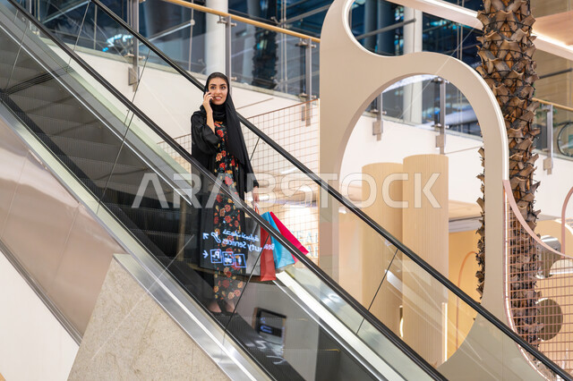 استخدام التقنية الحديثة, إجراء مكالمة هاتفية, الاستمتاع بإنتقاء المستلزمات والهدايا، الانتقال والنزول باستعمال السلم الكهربائي, إمرأة عربية خليجية اماراتية ترتدي الحجاب والعباءة تتجول في مركز التسوق تحمل الأكياس تستخدم الهاتف المحمول بإيماءات السعادة والسرور