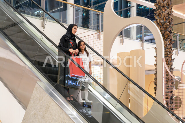الاستفادة من العروض والخصومات في المول, تعابير السعادة والسرور, الاستمتاع بالشراء وانتقاء المستلزمات والهدايا، الانتقال والنزول باستخدام السلم الكهربائي, إمرأة عربية خليجية اماراتية ترتدي الحجاب والعباءة تتجول في مركز التسوق برفقة ابنتها تحمل الأكياس الملونة