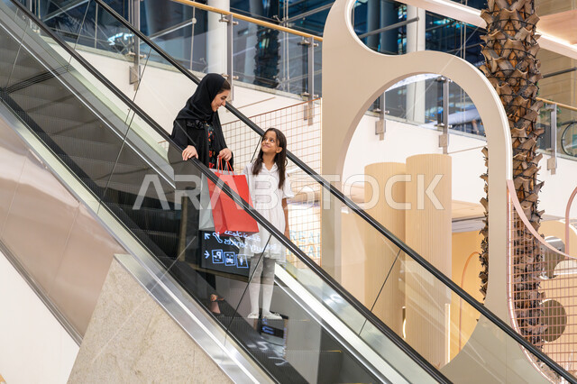 تعابير السعادة والسرور, الاستمتاع بالشراء وانتقاء المستلزمات والهدايا، الانتقال والنزول باستخدام السلم الكهربائي, إمرأة عربية خليجية اماراتية ترتدي الحجاب والعباءة تتجول في مركز التسوق برفقة ابنتها تحمل الأكياس الملونة، الاستفادة من العروض والخصومات في المول
