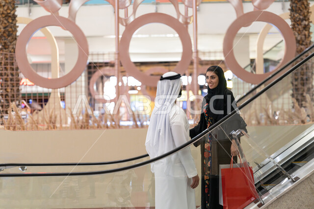 الانتقال والنزول والصعود باستخدام السلم الكهربائي, زوجين عربيين خليجيين اماراتيين يتجولون في مركز التسوق, إمرأة تقوم بحمل أكياس ورقية ملونة، الاستمتاع بالشراء وانتقاء المستلزمات والهدايا، الاستفادة من العروض والخصومات في المول التجاري، مفهوم الأسرة السعيدة