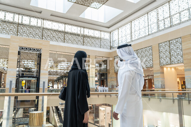 المشاركة في اختيارات التسوق وتبادل الآراء، قضاء وقت ممتع في إحدى المولات التجارية، الاستمتاع بالشراء والتبضع, صورة من الخلف لزوجين عربيين خليجيين اماراتيين يتجولان في المول، رجل يشير بإصبع السبابة الى شيء ما أو مكان معين, الإستفادة من العروض والتخفيضات الموسمية