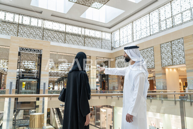 قضاء وقت ممتع في إحدى المولات التجارية، الاستمتاع بالشراء والتبضع, المشاركة في اختيارات التسوق وتبادل الآراء، صورة من الخلف لزوجين عربيين خليجيين اماراتيين يتجولان في المول، رجل يشير بإصبع السبابة الى شيء ما أو مكان معين, الإستفادة من العروض والتخفيضات الموسمية