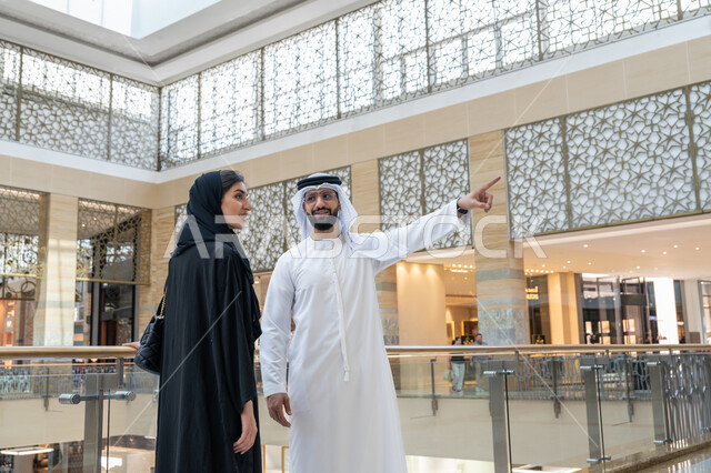 الاستمتاع بالشراء والتبضع, قضاء وقت ممتع في إحدى المولات التجارية، المشاركة في اختيارات التسوق وتبادل الآراء، زوجين عربيين خليجيين اماراتيين يتجولان في المول، رجل يشير بإصبع السبابة الى شيء ما أو مكان معين, الإستفادة من العروض والتخفيضات الموسمية