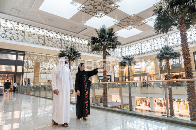 قضاء وقت ممتع في إحدى المولات التجارية، المشاركة في اختيارات التسوق وتبادل الآراء، زوجين عربيين خليجيين اماراتيين يتجولان في المول، إمرأة تشير بإصبع السبابة الى شيء ما أو مكان معين, الإستفادة من العروض والتخفيضات الموسمية، الاستمتاع بالشراء والتبضع