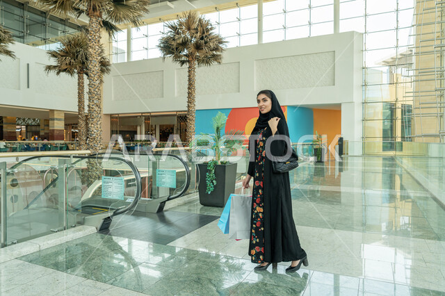 الاستمتاع بالشراء والتبضع, المتعة بالتسوق في إحدى المولات, إيماءات السعادة والسرور, الاستفادة من العروض والتخفيضات والحسومات الموسمية, إمرأة عربية خليجية اماراتية ترتدي الحجاب والعباءة السوداء تحمل بيدها الاكياس الورقية الملونة وتتجول في المول