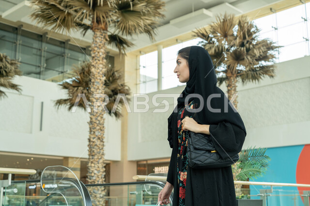 Enjoying shopping in a mall, gestures of happiness and pleasure, taking advantage of seasonal offers, discounts and reductions, enjoying shopping and shopping, a close-up of an Emirati Gulf Arab woman wearing a hijab and a black abaya, carrying bags in her hand and walking around the mall