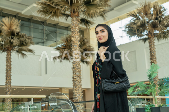 إيماءات السعادة والسرور, المتعة بالتسوق في إحدى المولات, الاستفادة من العروض والتخفيضات والحسومات الموسمية, الاستمتاع بالشراء والتبضع, صورة مقربة لإمرأة عربية خليجية اماراتية ترتدي الحجاب والعباءة السوداء وتتجول في المول, إقتناء المستلزمات اللازمة