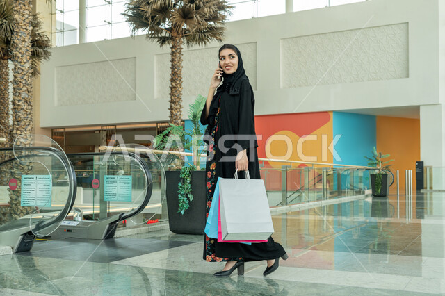 متعة التسوق في المول, استخدام التكنولوجيا والتقنية المتطورة الحديثة, إجراء مكالمة هاتفية, إمرأة عربية خليجية اماراتية ترتدي الحجاب والعباءة السوداء تحمل بيدها الأكياس الورقية الملونة وتستخدم الهاتف المحمول, الاستفادة من العروض والحسومات والتخفيضات الموسمية