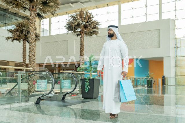 الاستمتاع  بالشراء والتبضع، متعة التسوق في إحدى المولات التجارية، الاستفادة من العروض والحسومات والتخفيضات الموسمية، رجل عربي خليجي اماراتي يرتدي الكندورة والغترة البيضاء والنظارات الطبية يتجول في المول ويحمل بيده الأكياس الورقية الملونة