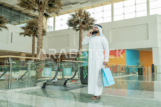 متعة التسوق في إحدى المولات، استخدام التكنولوجيا والتقنية الحديثة المتطورة، إجراء مكالمة هاتفية، رجل عربي خليجي اماراتي يرتدي الكندورة والغترة البيضاء والنظارات الطبية يمسك بيده الأكياس الورقية ويستخدم الهاتف المحمول، الاستفادة من العروض والحسومات الموسمية
