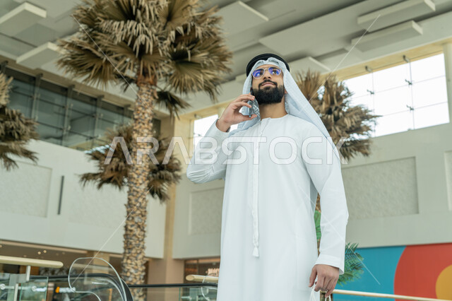 إجراء مكالمة هاتفية، استخدام التكنولوجيا والتقنية الحديثة المتطورة، صورة مقربة لرجل عربي خليجي اماراتي يرتدي الكندورة والغترة البيضاء والنظارات الطبية يمسك بيده الأكياس ويستخدم الهاتف المحمول، متعة التسوق في إحدى المولات، الاستفادة من العروض والحسومات الموسمية