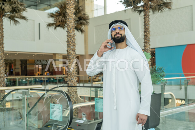 استخدام التكنولوجيا والتقنية الحديثة المتطورة، إجراء مكالمة هاتفية، صورة مقربة لرجل عربي خليجي اماراتي يرتدي الكندورة والغترة البيضاء والنظارات الطبية يمسك بيده الأكياس ويستخدم الهاتف المحمول، متعة التسوق في إحدى المولات، الاستفادة من العروض والحسومات الموسمية