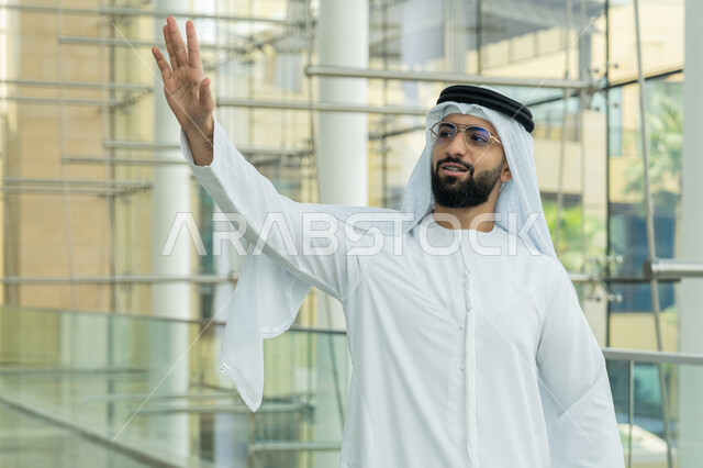 Having a nice time shopping in a mall, taking advantage of seasonal offers, discounts and sales, enjoying shopping, a close-up of an Emirati Gulf Arab man wearing a kandura, a white ghutra and eyeglasses, and pointing his hand upwards as a sign of greeting with expressions of happiness and pleasure