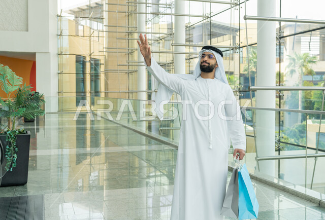 إقتناء المستلزمات اللازمة، قضاء وقع ممتع بالتسوق في إحدى المولات، الاستفادة من العروض والحسومات والتخفيضات الموسمية، الاستمتاع بالشراء والتبضع، رجل عربي خليجي اماراتي يرتدي الكندورة والغترة البيضاء والنظارات الطبية يمسك أكياس ويشيربيده للأعلى دلالة على التحية