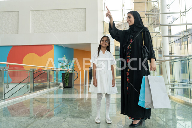 Participating in shopping choices and exchanging opinions, having a good time in a mall, enjoying shopping, an Emirati Gulf Arab woman wearing a hijab and abaya standing with her daughter holding bags in her hand and pointing with her index finger at something, taking advantage of seasonal offers and discounts