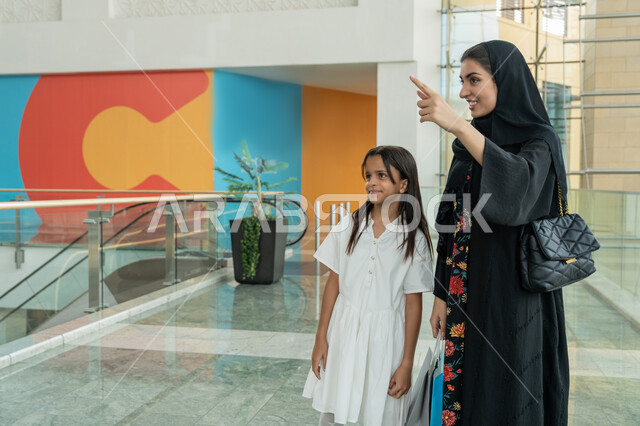قضاء وقت ممتع في إحدى المولات، الاستمتاع بالشراء والتبضع، المشاركة في اختيارات التسوق وتبادل الآراء، إمرأة عربية خليجية اماراتية ترتدي الحجاب والعباءة  تقف برفقة ابنتها تمسك بيدها الأكياس وتشير بإصبع السبابة لشيء ما، الإستفادة من العروض والتخفيضات الموسمية