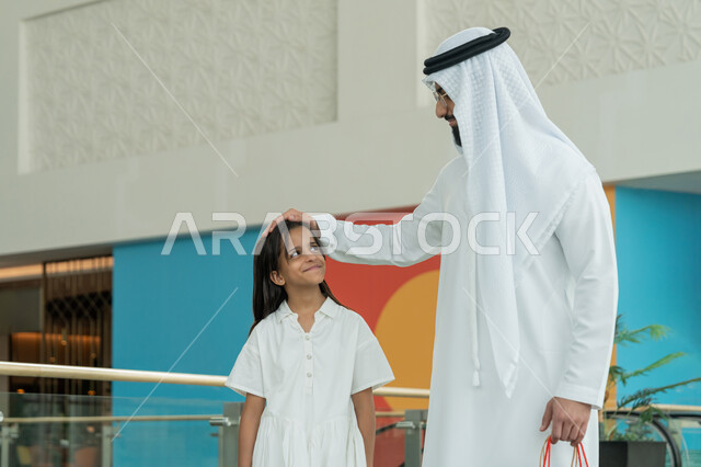 Enjoying shopping, participating in shopping choices and exchanging opinions, spending a good time in a shopping mall, an Arab Gulf Emirati man wearing a white kandura and ghutra standing with his daughter and placing his hand on her head with gestures of affection and love, taking advantage of seasonal offers and discounts