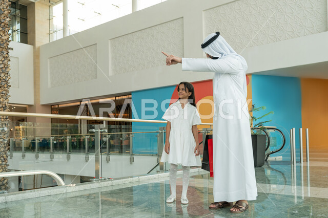 الإستفادة من العروض والتخفيضات الموسمية، المشاركة في اختيارات التسوق وتبادل الآراء، قضاء وقت ممتع في إحدى المولات التجارية، رجل عربي خليجي اماراتي يرتدي الكندورة والغترة البيضاء يقف برفقة ابنته ويشير بإصبع السبابة الى شيء ما، الاستمتاع بالشراء والتبضع في المول