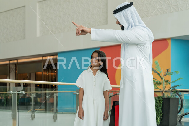 Participating in shopping choices and exchanging opinions, having a good time in a shopping mall, an Arab Gulf Emirati man wearing a white kandura and ghutra standing with his daughter and pointing at something with his index finger, taking advantage of seasonal offers and discounts, enjoying shopping in the mall