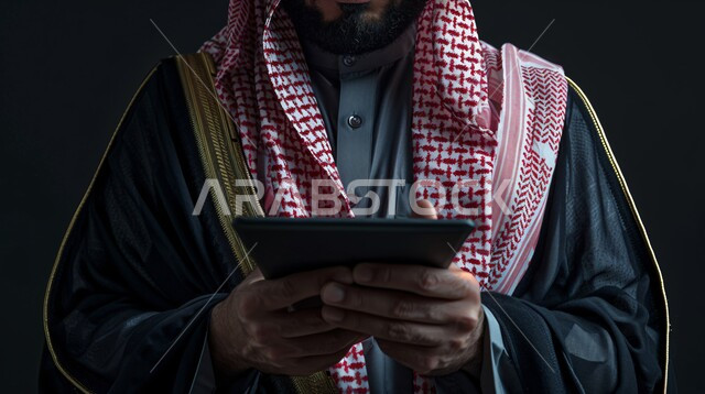 إدارة الأعمال عن بعد، استخدام الاجهزة التقنية الحديثة، الاهتمام بالأناقة والمظهر الخارجي، بورتريه مقرب لرجل عربي خليجي سعودي يرتدي الشماغ والثوب التقليدي باللون الاسود يحمل بيده الجهاز اللوحي، خلفية سوداء