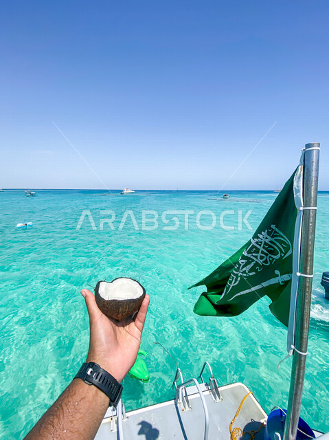 صورة ليد شاب سعودي يحمل بيده جوز الهند ، جزيره بياضه في جدة بالمملكة العربية السعودية، التسليه والترفيه ، الواجهة البحرية الطبيعية الخلابة ، علم السعودية على القارب، السياحة بالسعودية ، يخت داخل البحر