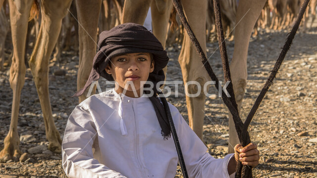 صورة مقربة لولد عماني يرتدي زي العماني التقليدي، يلتقط صورة بخلفية من الجمال بإيماءات وجه ويدين مختلفة، مزرعة تربية الإبل، فعاليات وأنشطة ترفيهيه بسلطنة عمان 
