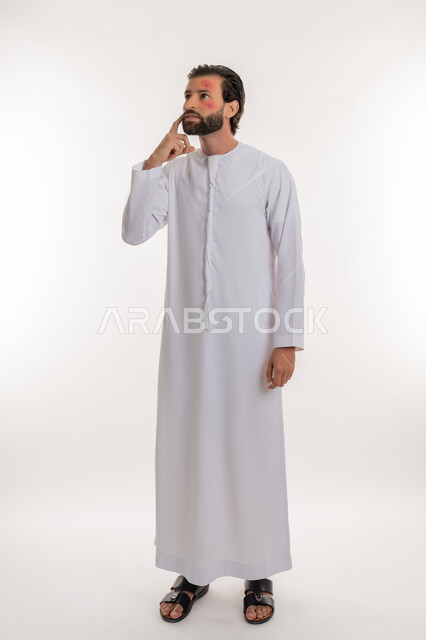 Need to visit a doctor and receive treatment, skin rash and infectious diseases, portrait of a young Arab Gulf Emirati man wearing a kandura pointing to the location of the disease, health problems and skin irritations, pimples spread on the skin of the face, skin infection and hypersensitivity, full body image, white background