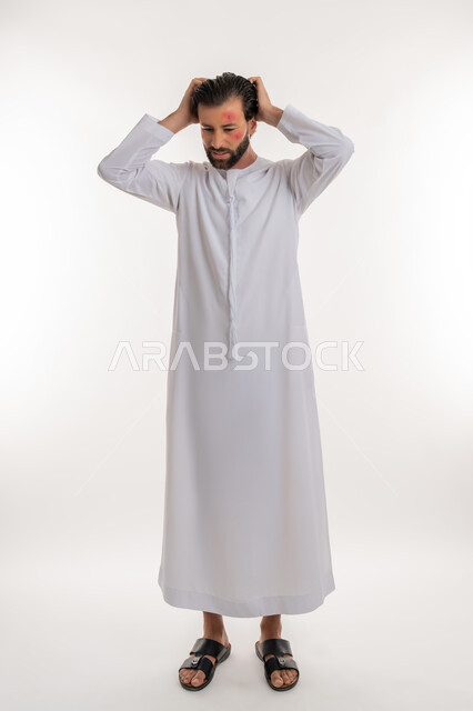 Gestures of stress and discomfort, rash and infectious diseases, portrait of a young Arab Gulf Emirati man wearing a kandura, putting his hands on his head with an expression of anxiety, health problems and skin irritations, pimples spread on the skin of the face, skin infection and hypersensitivity, full body image, white background