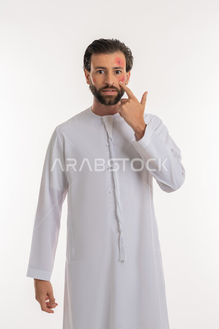 بثور منتشرة على بشرة الوجه, الإشارة إلى مكان الإصابة, بورتريه لرجل عربي خليجي اماراتي يرتدي الكندورة ينظر الى الكاميرا بإيماءات الإنزعاج والتعب, الإصابة بالحساسية الجلدية, المشاكل الصحية والتهجيات، الشعور بالأوجاع والالام, خلفية بيضاء