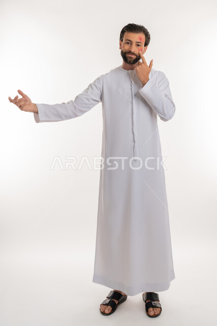 عدم المعرفة بامر ما, الإشارة إلى مكان المرض, بثور منتشرة على بشرة الوجه, بورتريه لرجل عربي خليجي اماراتي يرتدي الكندورة يرفع يده بايماءات الجهل, الإصابة بالحساسية الجلدية, المشاكل الصحية والتهجيات، صورة لكامل الجسم، خلفية بيضاء
