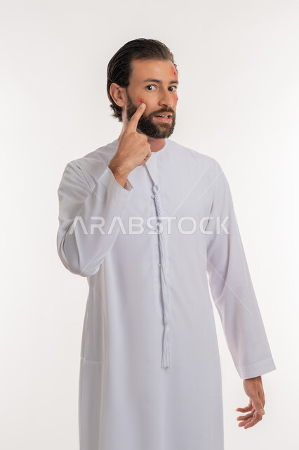 بثور منتشرة على بشرة الوجه, الإشارة إلى مكان الإصابة, بورتريه لرجل عربي خليجي اماراتي يرتدي الكندورة ينظر الى الكاميرا بإيماءات الإنزعاج والتعب, الإصابة بالحساسية الجلدية, المشاكل الصحية والتهجيات، الشعور بالأوجاع والالام, خلفية بيضاء