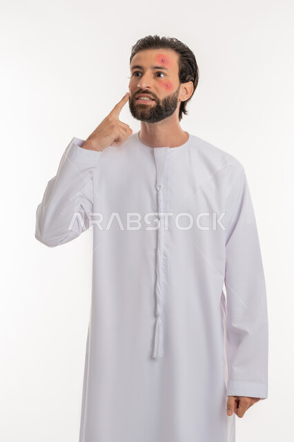 الطفح الجلدي والأمراض المعدية, الحاجة لزيارة الطبيب وتلقي العلاج, بورتريه لشاب عربي خليجي اماراتي يرتدي الكندورة يشير لمكان المرض, المشاكل الصحية وتهيجات الجلد, بثور منتشرة على بشرة الوجه, الإصابة الجلدية وفرط التحسس, خلفية بيضاء