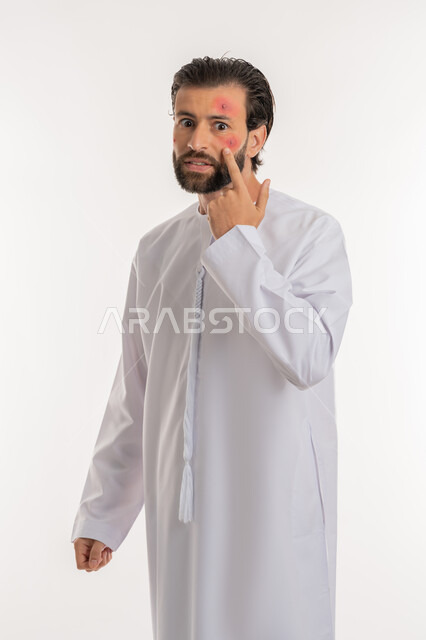 Skin allergy, pimples on face, pointing to the location of the lesion, side portrait of an Arab Gulf Emirati man wearing a kandura looking at the camera with gestures of annoyance and fatigue, health problems and spellings, feeling aches and pains, white background