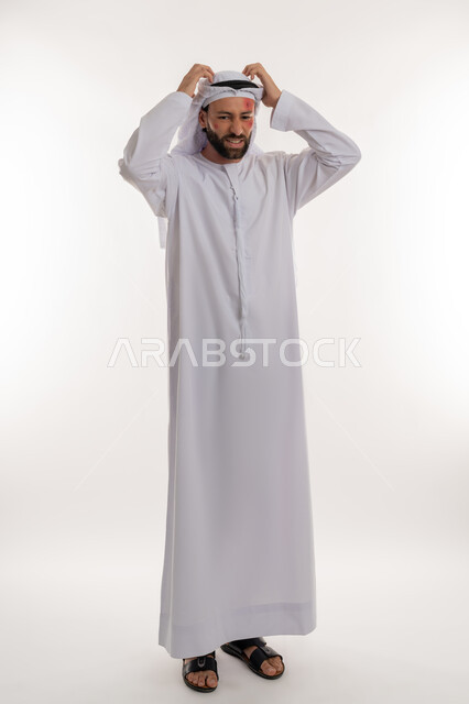 بثور منتشرة على بشرة الوجه, الأمراض المعدية والطفح الجلدي, فرط التحسس والإصابة الجلدية, الحاجة لزيارة الطبيب وتلقي العلاج, بورتريه لشاب عربي خليجي اماراتي يرتدي الكندورة والغترة يقوم بحك رأسه بإيماءات الإنزعاج, صورة لكامل الجسم، خلفية بيضاء