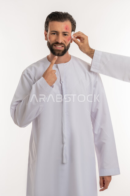 معالجة الأمراض والتهجيات، الحاجة لطلب المساعدة وزيارة الطبيب، فرط التحسس والإصابة الجلدية، بثور منتشرة على بشرة الوجه, بورتريه لرجل عربي خليجي اماراتي يرتدي الكندورة يقوم بالإشارة إلى مكان الإصابة، خلفية بيضاء