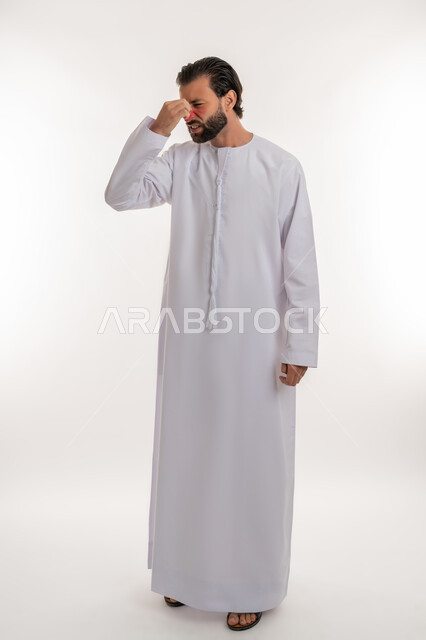 الشعور بالمرض وألم الرأس, الحاجة إلى العلاج والمساعده, بورتريه لرجل عربي خليجي اماراتي يرتدي الكندورة يضع يده على رأسه بإيماءات الانزعاج والتعب, آلام الشقيقة والصداع النصفي, صورة لكامل الجسم، خلفية بيضاء