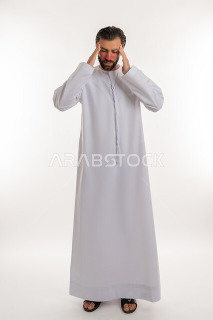 الحاجة إلى العلاج والمساعده, بورتريه لرجل عربي خليجي اماراتي يرتدي الكندورة يضع يديه على رأسه بإيماءات الانزعاج والتعب, الشعور بالمرض وألم الرأس, آلام الشقيقة والصداع النصفي, صورة لكامل الجسم، خلفية بيضاء