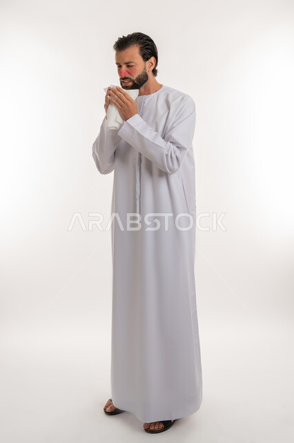 Using tissues to sneeze, allergic rhinitis, feeling sick and having a cold and needing treatment and medication, side portrait of a young Emirati Gulf Arab man wearing a kandura raising his hands to his face with sneezing gestures, full body image, white background