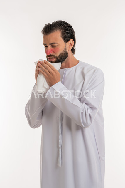 الشعور بالمرض و الزكام والحاجة الى العلاج والدواء, التهاب الأنف التحسسي, استخدام المناديل الورقيه للعطس, بورتريه من الجانب لشاب عربي خليجي اماراتي يرتدي الكندورة يرفع يديه على وجهه بإيماءات العطاس, خلفية بيضاء