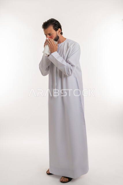 استخدام المناديل الورقيه للعطس, التهاب الأنف التحسسي, الشعور بالمرض و الزكام والحاجة الى العلاج والدواء, بورتريه من الجانب لشاب عربي خليجي اماراتي يرتدي الكندورة يرفع يديه على وجهه بإيماءات العطاس, صورة لكامل الجسم، خلفية بيضاء