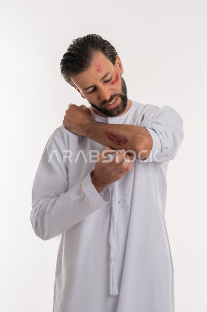 الحاجة الى الرعاية الطبية المنزلية، اصابات وكدمات جسدية، التعرض للعنف والاذى، الشعور بالالم الشديد، بورتريه لرجل عربي خليجي اماراتي مصاب يرتدي الكندورة يقوم بالنظر الى الجروح بتعابير التعب والارهاق، خلفية بيضاء