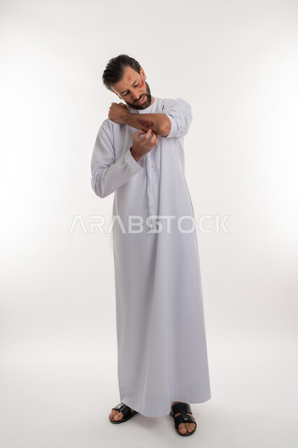 الشعور بالالم الشديد، التعرض للعنف والاذى، اصابات وكدمات جسدية، الحاجة الى الرعاية الطبية المنزلية، بورتريه لرجل عربي خليجي اماراتي مصاب يرتدي الكندورة يقوم بالنظر الى الجروح بتعابير التعب والارهاق، صورة لكامل الجسم، خلفية بيضاء