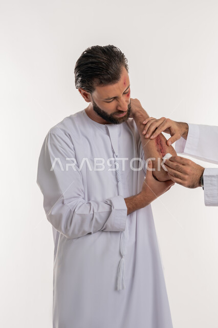 تلقي الرعاية الطبية المنزلية، اصابات وكدمات جسدية، التعرض للعنف والاذى، الشعور بالالم الشديد، بورتريه لرجل عربي خليجي اماراتي مصاب يرتدي الكندورة يقوم بالنظر الى الجروح بتعابير التعب والارهاق، خلفية بيضاء