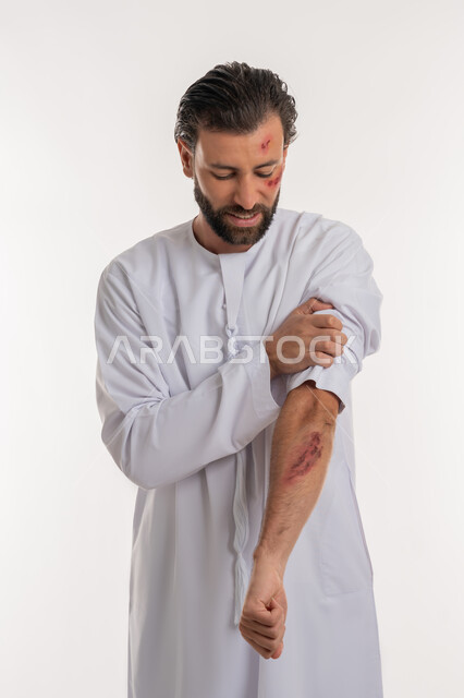 اصابات وكدمات جسدية، التعرض للعنف والاذى، الشعور بالالم الشديد، الحاجة الى الرعاية الطبية المنزلية، بورتريه لرجل عربي خليجي اماراتي مصاب يرتدي الكندورة يقوم بالنظر الى الجروح بتعابير التعب والارهاق، خلفية بيضاء