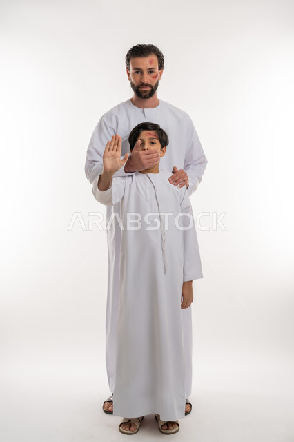 الغضب والسلوك العدواني, المشاكل والخلافات الاسرية، اصابات و جروح جسدية، بورتريه لولد عربي خليجي اماراتي يرتدي الكندورة يتعرض للعنف من قبل والده يرفع يده بايماءات الرفض والامتناع، تعابير الآلم والحزن، قضايا اجتماعيه أسريه، صورة لكامل الجسم، خلفية بيضاء